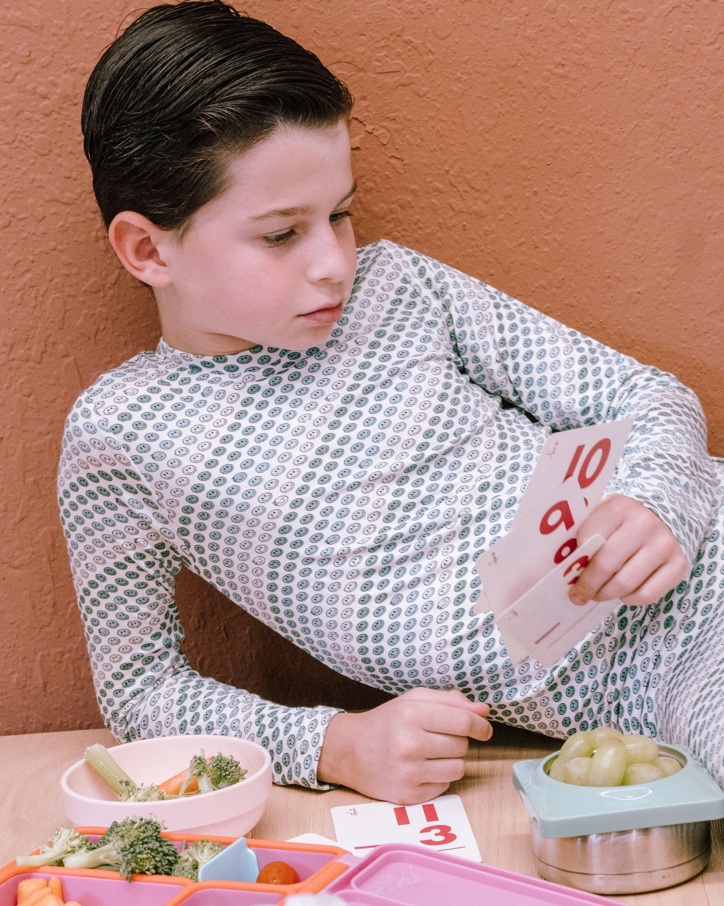 A young boy with dark hair sits at a table against a textured terracotta wall, wearing a long-sleeve white shirt with a gray geometric pattern. He is holding a card with red numbers, with colorful bento boxes and food bowls on the table in front of him.
