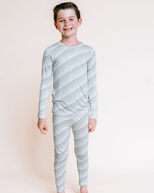 A young boy stands barefoot against a plain light beige studio background, wearing a long-sleeve pajama set with a repeating diagonal geometric pattern in light blue and white.