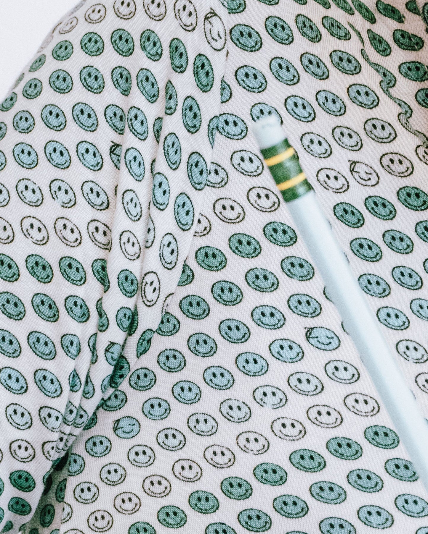 A close-up lifestyle shot of a person wearing a long-sleeve button-up shirt with a green, white, and gray smiley face pattern, writing on a piece of paper at a desk.