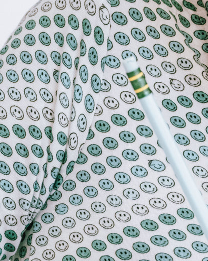 A close-up lifestyle shot of a person wearing a long-sleeve button-up shirt with a green, white, and gray smiley face pattern, writing on a piece of paper at a desk.
