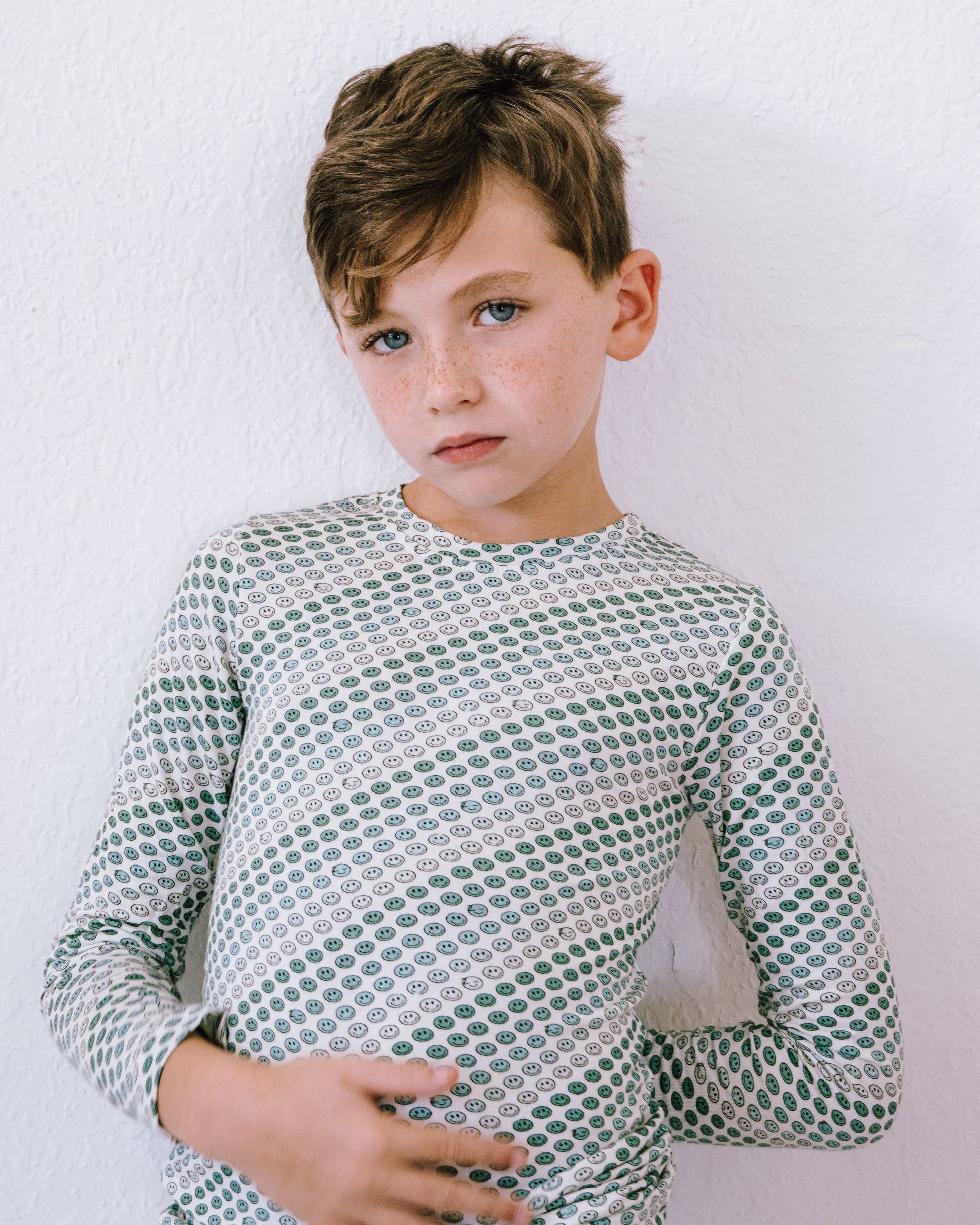 A young boy with light brown hair wearing a green and white patterned long-sleeve shirt, standing against a white textured wall.