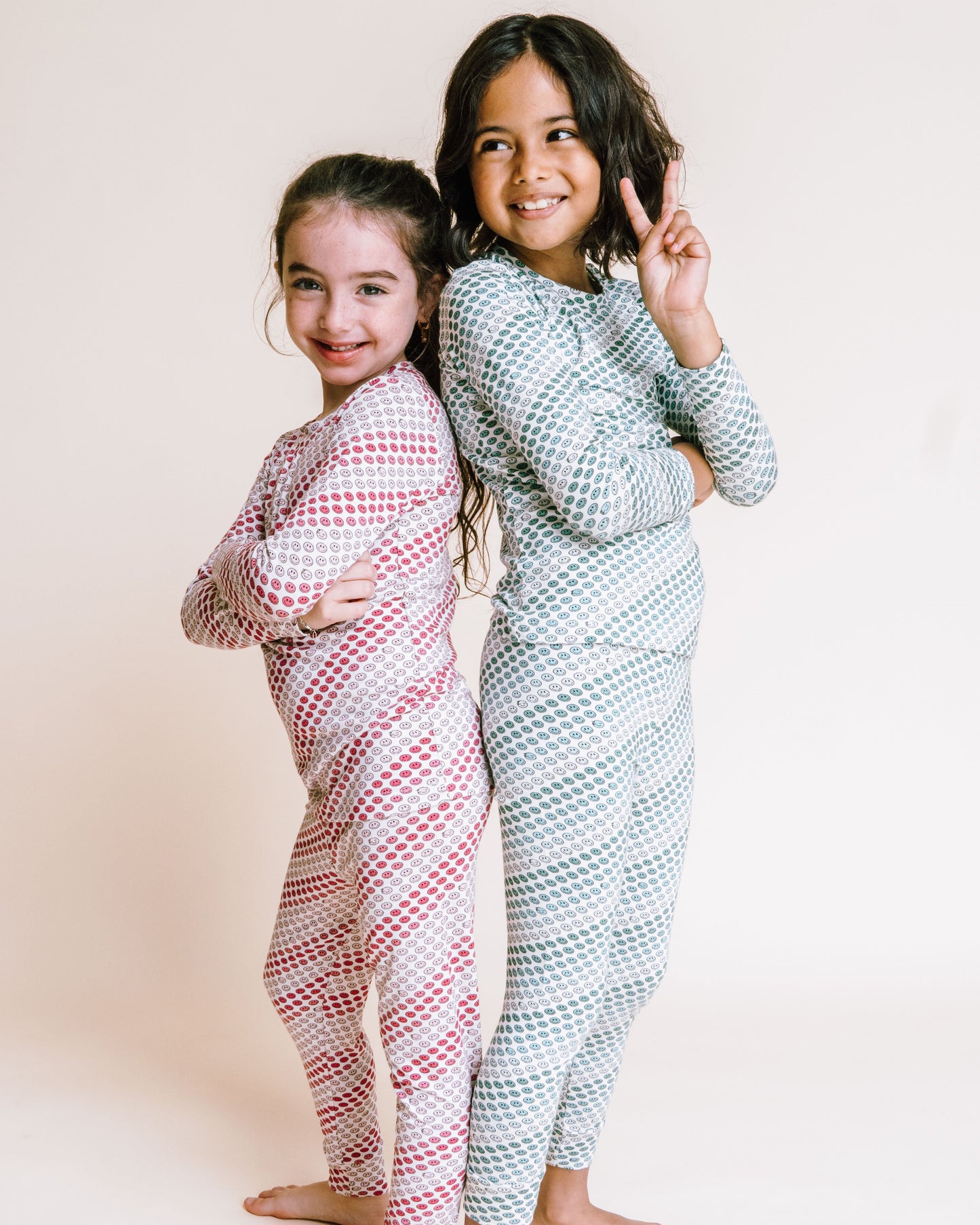 Two young girls standing barefoot against a plain cream background, wearing matching long-sleeve patterned pajamas. The girl on the left wears a pink and white diagonal striped set, and the girl on the right wears a light blue and white diagonal striped set.
