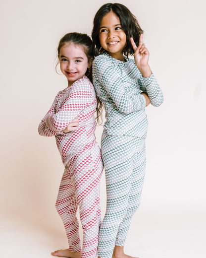 Two young girls standing barefoot against a plain cream background, wearing matching long-sleeve patterned pajamas. The girl on the left wears a pink and white diagonal striped set, and the girl on the right wears a light blue and white diagonal striped set.