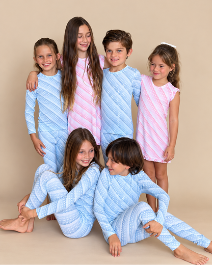 Six children posing together in matching patterned pajamas against a solid beige studio background. Three children wear light blue and white textured long-sleeve pajama sets, while three wear light pink and white textured long-sleeve pajama dresses.