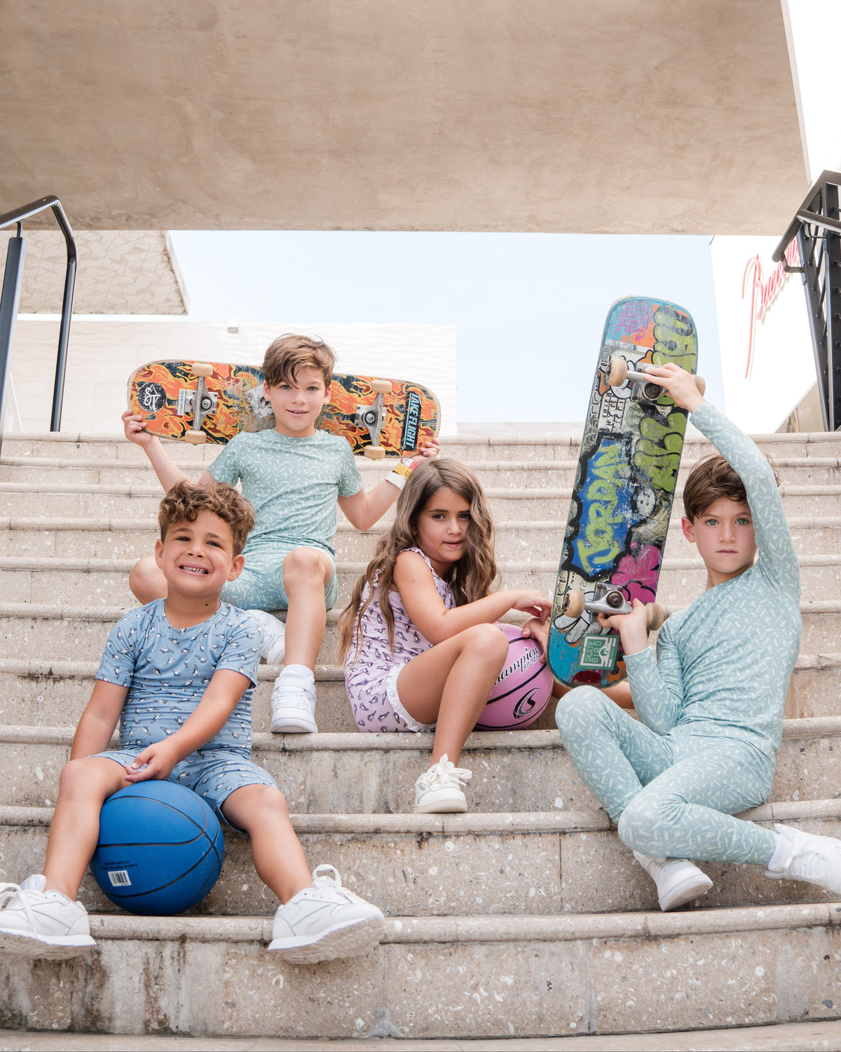 Five children sitting on outdoor concrete stairs under a bridge, wearing matching light blue and lavender athletic sets and holding colorful skateboards and a blue basketball.