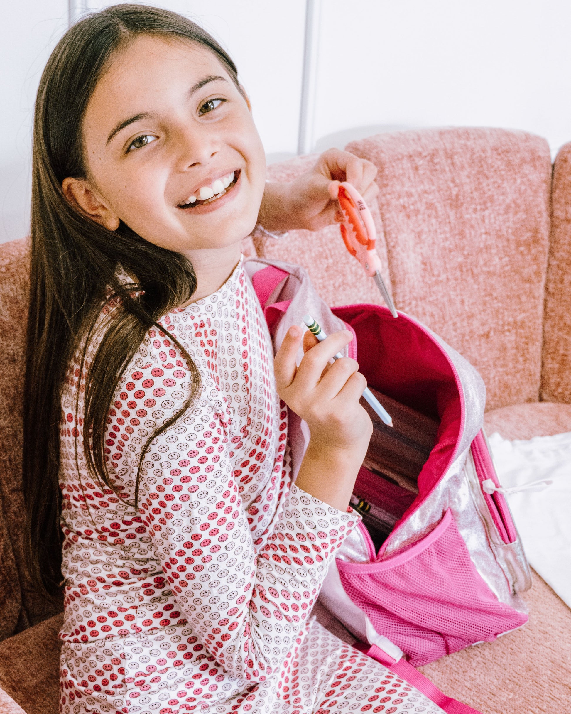A young girl with long brown hair smiles while sitting on a pink couch, wearing a pink and white patterned pajama set. She is holding a pink backpack and a small orange item.