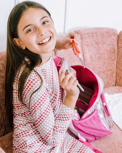 A young girl with long brown hair smiles while sitting on a pink couch, wearing a pink and white patterned pajama set. She is holding a pink backpack and a small orange item.