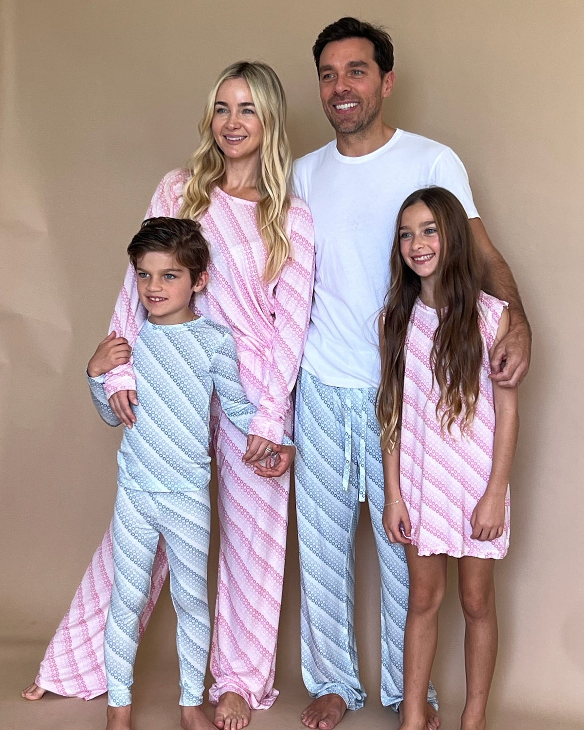 A family of four posing together in matching patterned pajamas. The mother and daughter wear pink and light blue striped pajama sets, while the father wears a white t-shirt with matching light blue pajama pants, and the son wears a blue and white striped pajama set.