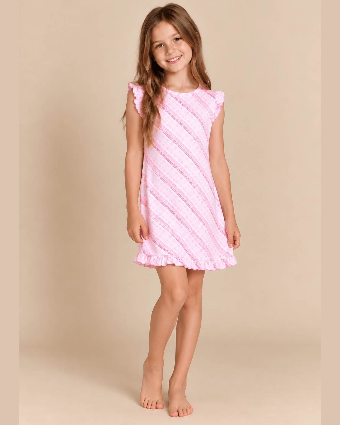 A young girl with long light brown hair stands barefoot against a solid beige studio background, wearing a sleeveless pink dress with diagonal textured stripes and ruffled trim.