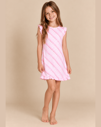 A young girl with long light brown hair stands barefoot against a solid beige studio background, wearing a sleeveless pink dress with diagonal textured stripes and ruffled trim.