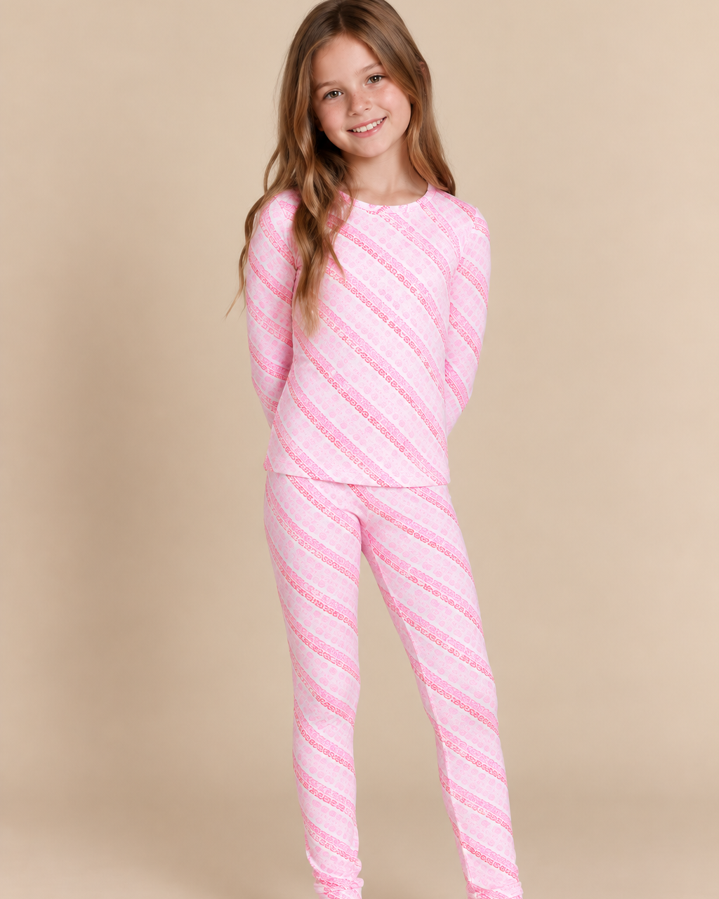 A young girl with long light brown hair stands barefoot against a solid beige studio background, wearing a matching two-piece pajama set with diagonal pink and white stripes.