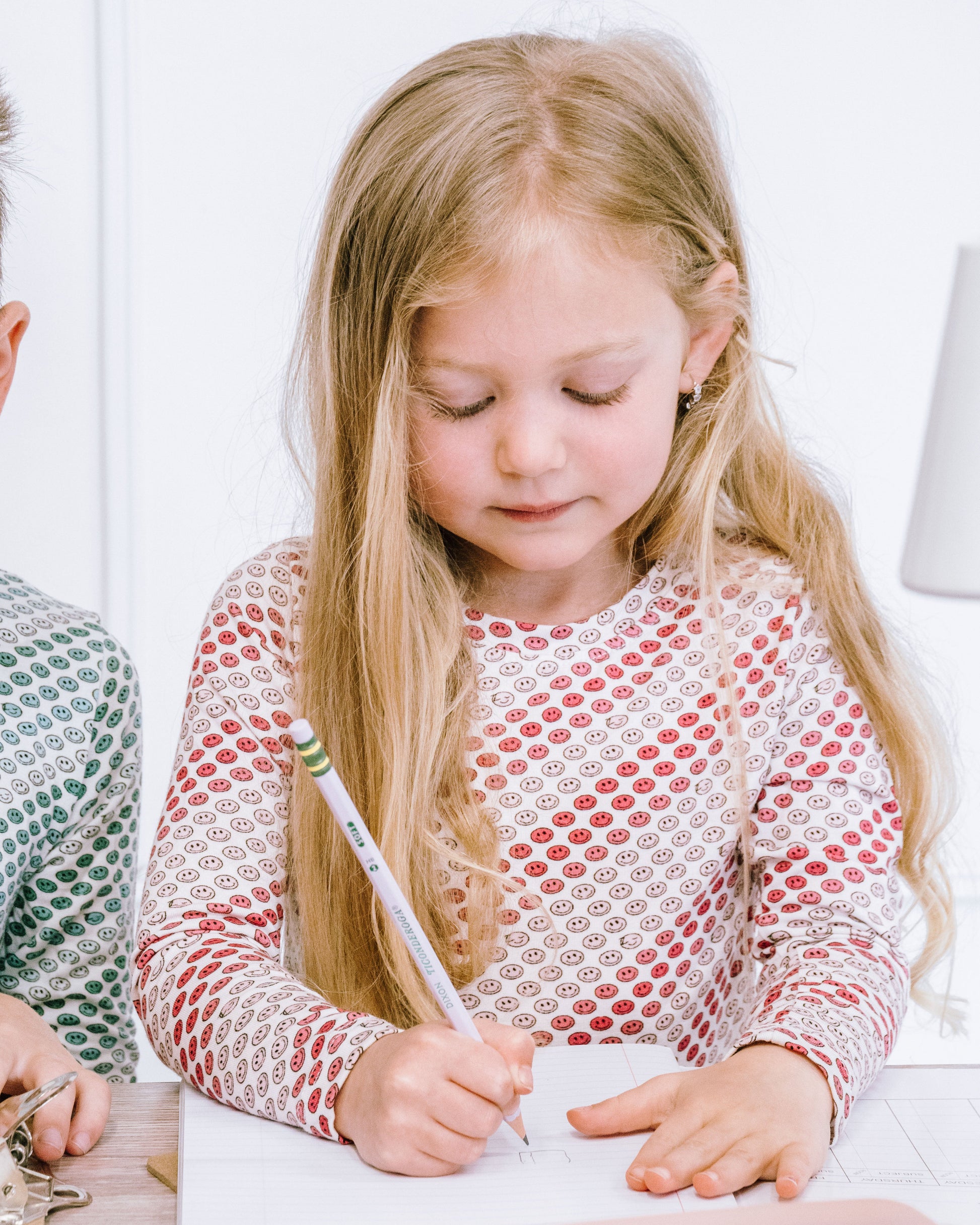 A young girl with long blonde hair sits at a table, writing on a piece of paper with a pencil. She is wearing a long-sleeve white top with a red and gray scalloped pattern.