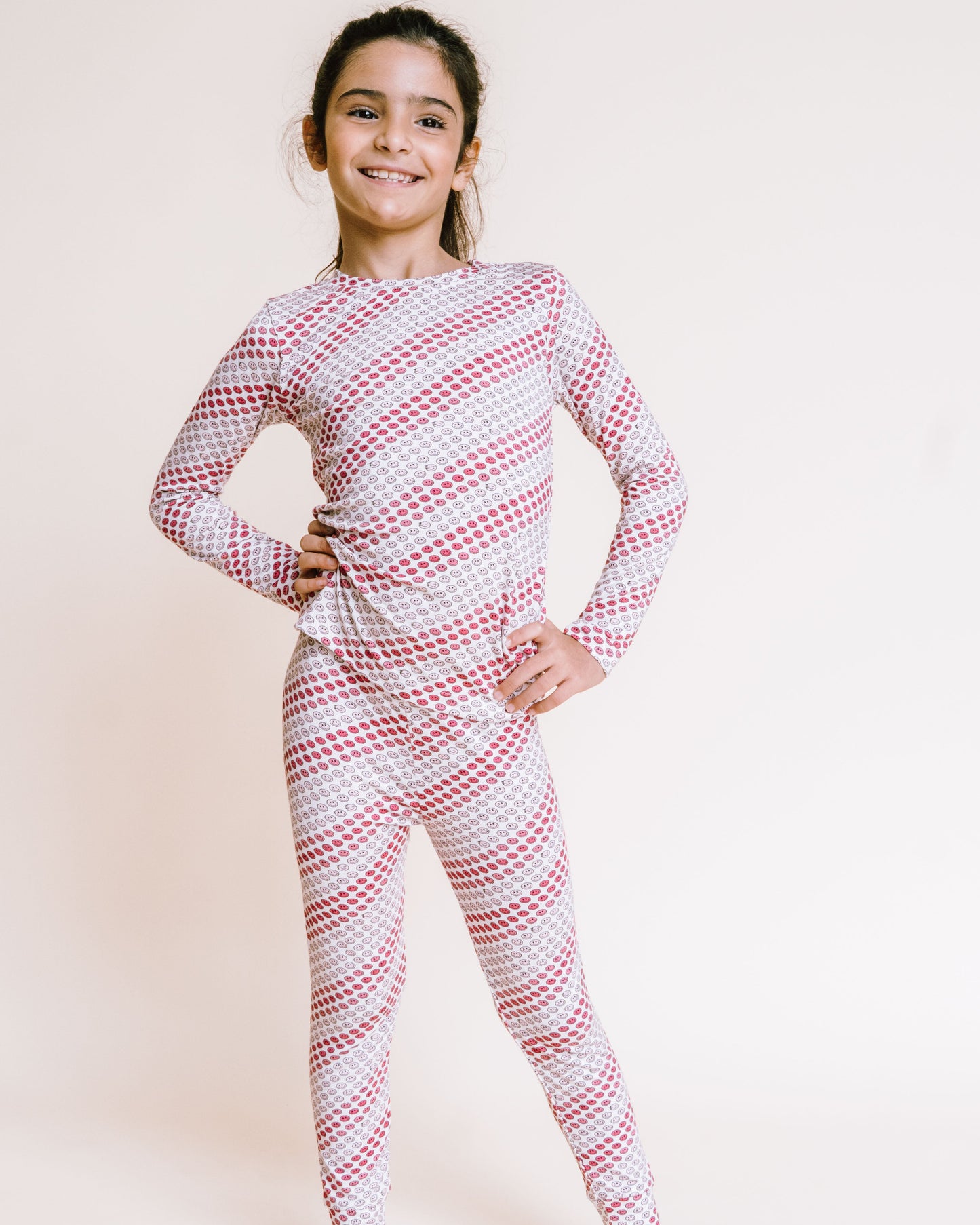 A young girl stands barefoot against a plain light beige studio background, wearing a long-sleeve one-piece pajama set with a red and white diagonal stripe pattern.