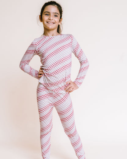 A young girl stands barefoot against a plain light beige studio background, wearing a long-sleeve one-piece pajama set with a red and white diagonal stripe pattern.